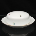Porcelana Renner, Porto Alegre - RS (Fundada em 1937). Manteigueira em porcelana nacional, composta por prato com receptáculo central e tampa de perfil abaulado. Esmaltagem vítrea na cor branca, decoração policroma com pequenos ramos florais esparsos em azul, rosa, violeta, amarelo e verde; filetagem dourada contornando a tampa e a borda do prato. Marca da manufatura no verso. Medidas: Alt.: 6,5 x Diam.: 17,5 cm.