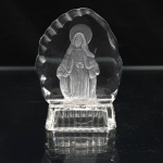 Escultura devocional executada em cristal incolor moldado, representando Nossa Senhora das Graças, de pé, com véu, túnica longa cingida à cintura, manto e mãos estendidas em gesto característico da iconografia mariana. Figura executada em acabamento fosco, contrastando com o corpo transparente da peça; auréola circular e contorno externo irregular, assente sobre base retangular moldada. Medidas: Alt.: 10 x Comp.: 7,5 x Larg.: 3,5 cm.