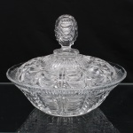 Bomboniere com tampa em vidro prensado translúcido de alta densidade, manufaturada na Boêmia, antiga Tchecoslováquia, atribuída à produção de Josef Inwald, circa 1930. Decoração de estética Art Déco exibe bojo aproximadamente obcônico moldado com faixas radiais de arcos crescentes sobrepostos em relevo côncavo-convexo, tampa abaulada apresenta remate por pega com lapidação facetada e arestas serrilhadas nas bordas, fundo preenchido por medalhão em estrela de múltiplos raios (starburst) obtido por compressão mecânica durante o processo de fundição. Medidas: Alt.: 19,5 x Diam.: 25 cm.