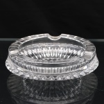 Cinzeiro circular em cristal prensado, diáfano e incolor, de paredes espessas, com borda facetada provida de quatro apoios semicirculares para cigarros. Corpo trabalhado por decoração geométrica, apresentando sequência de motivos lanceolados dispostos radialmente e faixa periférica de facetas. Medidas: Alt.: 4 x Diam.: 15 cm.