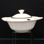 W.H. GRINDLEY, Staffordshire - Inglaterra(Fundada por William Harry Grindley em 1880). Sopeira em faiança inglesa, padrão "CREAMPETAL", c.1940-50. Esmaltagem vítrea de tonalidade marfim claro, bojo hemisférico, orla modelada com faixa perimetral ondulada, percorrida por perolados relevados na própria pasta, ornamentada por filetagem à ouro contornando a borda e a caldeira, abas laterais integradas e decoradas com ramagens, tampa abaulada encimada por pomo em forma de fruto, dotada de abertura lateral para passagem da concha. Marca da manufatura no verso. Medidas: Alt.: 17 x Diam.: 30 cm.