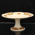 Wood & Sons, Staffordshire - Inglaterra (fundada em 1865/ ativa até 2005). Doceira de pé alto em faiança inglesa, padrão "Woods Ivory Ware", Inglaterra, circa 1930-1940. Prato circular de bordo ondulado e canelado, elevado sobre coluna central de perfil afunilado, terminada em base circular moldada. Ornamentação por decalcomania policroma composta por ramalhetes florais e cartelas de desenho geometrizado, distribuídos em sequência pela aba e repetidos junto à base, nas tonalidades laranja, ocre, preto e creme, filete preto acompanha a delimitação interna da aba. Marca de fábrica "Woods Ivory Ware  England", com coroa, aposta sob a base. Medidas: Alt.: x Diam.: cm.