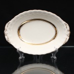 W.H. GRINDLEY, Staffordshire - Inglaterra(Fundada por William Harry Grindley em 1880). Travessa em faiança inglesa, padrão "CREAMPETAL", c.1940-50. Esmaltagem vítrea de tonalidade marfim claro, formato oval de interior ligeiramente côncavo, orla modelada com faixa perimetral ondulada, percorrida por perolados relevados na própria pasta, ornamentada por filetagem à ouro contornando a borda e a caldeira, abas laterais integradas e decoradas com ramagens. Apresenta ínfimos desgastes na douradura. Marca da manufatura no verso. Medidas: Alt.: 4 x Comp.: 21 x Larg.: 15 cm.
