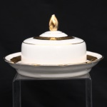 Porcelana Rio Branco / Cia. Brasileira de Vidros, Rio de Janeiro - RJ (fundada em 1947, ativa até 1967). Manteigueira em porcelana nacional, composta por prato, recipiente interno e tampa em campânula, rematada por pomo em forma de botão floral. Esmaltagem vítrea na cor branca, com bordos de perfil ondulado. Ornamentação constituída por faixa dourada com friso de ramagens florais estilizadas, complementada por filetes a ouro no bordo e na tampa. Marca da mnanufatura ao fundo. Medidas: Alt.: 8 x Diam.:  15cm.