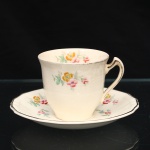 Alfred Meakin, Staffordshire - Inglaterra(Fundada em 1875, ativa até 1976). Xícara para café em faiança inglesa, padrão "Marigold", circa 1930-1940. Xícara de corpo obcônico e alça estilizada de perfil anguloso, acompanhada de pires circular com bordo recortado. Esmlatação de tom marfim dotada de decoração floral policroma composta por pequenos ramos distribuídos pela superfície, em tonalidades de rosa, laranja, azul e verde, complementada por filetação em douradura no bordos e na alça. No fundo da xícara, marca de manufatura "Alfred Meakin  Marigold". Apresenta craquelê do vidrado e desgaste da filetação. Medidas: Xícara: Alt.: 6 x Comp.: 8 x Larg.: 6,5cm; Medida pires: Diam.: 12cm.