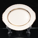 W.H. GRINDLEY, Staffordshire - Inglaterra(Fundada por William Harry Grindley em 1880). Travessa rasa em faiança inglesa, padrão "CREAMPETAL", c.1940-50. Esmaltagem vítrea de tonalidade marfim claro, formato oval, orla modelada com faixa perimetral ondulada, percorrida por perolados relevados na própria pasta, ornamentada por filetagem à ouro contornando a borda e a caldeira, abas laterais integradas e decoradas com ramagens. Apresenta ínfimos degastes de uso na douradura. Marca da manufatura no verso. Medidas: Comp.: 26,5 x Larg.: 21,5 cm.