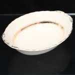 W.H. GRINDLEY, Staffordshire - Inglaterra(Fundada por William Harry Grindley em 1880). Travessa funda em faiança inglesa, padrão "CREAMPETAL", c.1940-50. Esmaltagem vítrea de tonalidade marfim claro, formato oval de interior côncavo, orla modelada com faixa perimetral ondulada, percorrida por perolados relevados na própria pasta, ornamentada por filetagem à ouro contornando a borda e a caldeira, abas laterais integradas e decoradas com ramagens. Apresenta ínfimas manchas do tempo. Marca da manufatura no verso. Medidas: Alt.: 7 x Comp.: 22,5 x Larg.: 18 cm.