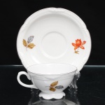 Porzellanfabrik Zeh, Scherzer & Co, Baviera - Alemanha (Fundada em 1880). Excepcional chávena para café em porcelana alemã da década de 1930, estilo e período Art Déco. Esmaltagem vítrea na cor branca, dotada de decoração em estampagem policromada, com flores e ramagens outonais nas tonalidades coral, ocre, azul e amarelo, realçada por filetação prateada nas bordas e detalhes. Bojo hemisférico assentado sobre base cirular elevada de borda recortada, asa lateral em voluta. Bordas da xícara e do pires possuem contorno relevado com sucessão de frisos radias interrompidos por folha de acanto estilizada. Marca da manufatura ao fundo, acompanhada de selo comemorativo de 50 anos de fundação da marca com a inscrição "50 JAHRE"(O timbre "50 Jahre" (50 anos) identifica a tiragem limitada produzida pela Porzellanfabrik Zeh, Scherzer & Co. AG de forma exclusiva no decorrer do ano de 1930, em celebração ao cinquentenário de fundação da fábrica em Rehau, na Baviera). Medidas: Xícara: Alt.: 4 x Comp.: 9 x Larg.: 7,5 cm; Pires: Diam.: 12 cm.