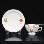 Porzellanfabrik Zeh, Scherzer & Co, Baviera - Alemanha (Fundada em 1880). Excepcional chávena para café em porcelana alemã da década de 1930, estilo e período Art Déco. Esmaltagem vítrea na cor branca, dotada de decoração em estampagem policromada, com flores e ramagens outonais nas tonalidades coral, ocre, azul e amarelo, realçada por filetação prateada nas bordas e detalhes. Bojo hemisférico assentado sobre base cirular elevada de borda recortada, asa lateral em voluta. Bordas da xícara e do pires possuem contorno relevado com sucessão de frisos radias interrompidos por folha de acanto estilizada. Marca da manufatura ao fundo, acompanhada de selo comemorativo de 50 anos de fundação da marca com a inscrição "50 JAHRE"(O timbre "50 Jahre" (50 anos) identifica a tiragem limitada produzida pela Porzellanfabrik Zeh, Scherzer & Co. AG de forma exclusiva no decorrer do ano de 1930, em celebração ao cinquentenário de fundação da fábrica em Rehau, na Baviera). Medidas: Xícara: Alt.: 4 x Comp.: 9 x Larg.: 7,5 cm; Pires: Diam.: 12 cm.