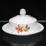 Porzellanfabrik Zeh, Scherzer & Co, Baviera - Alemanha (Fundada em 1880). Excepcional manteigueira em porcelana alemã da década de 1930, estilo e período Art Déco. Esmaltagem vítrea na cor branca, dotada de decoração em estampagem policromada, com flores e ramagens outonais nas tonalidades coral, ocre, azul e amarelo, realçada por filetação prateada nas bordas e detalhes. Tampa circular de topo abaulado, encimado por pomo com feitio de fruto, prato dotado de receptáculo cilíndrico central integrado. Marca da manufatura ao fundo, acompanhada de selo comemorativo de 50 anos de fundação da marca com a inscrição "50 JAHRE"(O timbre "50 Jahre" (50 anos) identifica a tiragem limitada produzida pela Porzellanfabrik Zeh, Scherzer & Co. AG de forma exclusiva no decorrer do ano de 1930, em celebração ao cinquentenário de fundação da fábrica em Rehau, na Baviera). Medidas: Alt.: 10,5 x Diam.: 19cm.