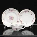 CHODZIEZ PORCELAIN, Chodziez - Polônia(Fundada em 1852 ou 1854, por Hermann Müller e Ludwik Schnorr). Belíssimo trio para chá em porcelana polonesa da década de 1940; esmaltagem vítrea na cor branca, bojo hemisférico assentado sobre base circular recortada, alça lateral em voluta; decorada com padrão floral em estampagem policromada, composto de arranjo floral nas tonalidades rosa, azul, amarelo e verde, e buquês distribuídos nas orlas do pires e do prato. Apresenta filetagem à ouro nas bordas interna e externa. Orla delimitada por barrado contínuo relevado na própria pasta. Padrão decorativo integralmente replicado no pires e no prato. Marca da manufatura no verso. Medidas: Xícara: Alt.: 5 x Comp.: 12 x Larg.: 10 cm; Prato: Diam.: 17 cm; Pires: Diam.: 15 cm.