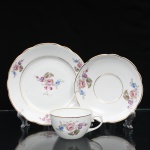 Porcelana Real, Mauá - SP(Fundada em 1943 por Fritz Erwin Schmidt). Trio para chá composto por xícara, pires e prato de sobremesa em porcelana de pasta, fabricação nacional, circa 1960-1970). Esmaltagem vítrea na cor branca, bojo hemisférico, asa lateral escultural em formato de voluta com terminação em rolo superior, ornamentação com motivos pictóricos policromados por processo de decalcomania fina inspirados no estilo Deutsche Blumen, completado com filetagem à ouro nas bordas e margens. Centro do prato de sobremesa e reservas periféricas dos componentes retratam um buquê clássico composto por rosa púrpura centralizada, violetas, miosótis e folhagens detalhadas em tons esmeralda, aba percorrida por gomos ondulares suaves. Marca da manufatura no verso. Medidas: Xícara: Medidas: Alt.: 5 x Comp.: 10,5 x Larg.: 8,5 cm; Prato: 18 cm; Pires: Diam.: 14,5 cm.