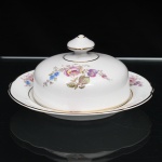 Porcelana Real, Mauá - SP(Fundada em 1943 por Fritz Erwin Schmidt). Belíssima manteigueira executada em porcelana nacional, circa 1960-1970. Esmaltagem vítrea na cor branca, ornamentação com motivos pictóricos policromados por processo de decalcomania fina inspirados no estilo "Deutsche Blumen", realçada por filetagem à ouro nas bordas e margens. Tampa em cúpula abobadada encimada por pomo facetado com seis lados, prato dotado de receptáculo cilíndrico central integrado, borda recortada, realçada por filetagem à ouro nas bordas e margens. Marca da manufatura ao fundo. Medidas: Alt.: 8 x Diam.: 15 cm.