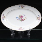Porcelana Real, Mauá - SP(Fundada em 1943 por Fritz Erwin Schmidt). Belíssima travessa oval executada em porcelana nacional, circa 1960-1970. Esmaltagem vítrea na cor branca, ornamentação com motivos pictóricos policromados por processo de decalcomania fina inspirados no estilo "Deutsche Blumen", replicado nas reservas periféricas da orla, aba percorrida por gomos ondulares suaves, realçado com filetagem à ouro nas bordas e margens. Marca da manufatura no verso. Medidas: Comp.: x Larg.: cm.