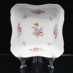 Porcelana Schmidt, Santa Catarina, Brasil (Fundada em 1945). Saladeira em porcelana nacional, c.1950/60, executado com elementos derivados do repertório Luís XV. Formato quandrangular de quinas arredondas, borda ondulada, revirada para fora, contornada por friso em douradura, orla circundada por faixa de motivo fitomorfo relevado na própria pasta. Esmaltagem vítrea na cor branca, decoração floral policroma por decalcomania, distribuída pelo fundo e pelas paredes internas em buquês e ramagens com rosas, tulipa e flores miúdas, em rosa, azul, vermelho, amarelo e verde. Marca da manufatura ao fundo. Medidas: Alt.: 7 x Comp.: 23 x Larg.: 23 cm.