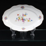 Porcelana Schmidt, Santa Catarina, Brasil (Fundada em 1945). Travessa rasa em porcelana nacional, c.1950/60, executado com elementos derivados do repertório Luís XV. Formato oval, borda ondulada contornada por friso em douradura, orla circundada por faixa de motvo fitomorfo relevado na própria pasta. Esmaltagem vítrea na cor branca, decoração floral policroma aplicada por decalcomania, constituída por ramagens e corolas em rosa, azul, vermelho, amarelo e folhagens verde. Marca da manufatura ao fundo. Medidas: Comp.: 32,5 x Larg.: 21 cm.