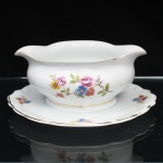 Porcelana Schmidt, Santa Catarina, Brasil (Fundada em 1945). Molheira em porcelana nacional, c.1950/60, executado com elementos derivados do repertório Luís XV. Bojo elipsoide dotado de vertdouros opostos, borda ondulada e revirada para fora, fixada sobre prato de apoio circular de bordo recortado, circundado por ramagens relevadas na própria pasta. Esmaltagem vítrea na cor branca, decoração floral policroma aplicada por decalcomania, constituída por ramagens e corolas em rosa, azul, vermelho, amarelo e folhagens verde, complementada por filetagem em dourada nas bordas e base. Marca da manufatura ao fundo. Medidas: Alt.: 8,5 x Comp.: 16,5 x Larg.: 14 cm; Prato de apoio: Diam.: 18,5 cm.