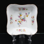 Porcelana Schmidt, Santa Catarina, Brasil (Fundada em 1945). Saladeira em porcelana nacional, c.1950/60, executado com elementos derivados do repertório Luís XV. Formato quandrangular de quinas arredondas, borda ondulada, revirada para fora, contornada por friso em douradura, orla circundada por faixa de motivo fitomorfo relevado na própria pasta. Esmaltagem vítrea na cor branca, decoração floral policroma aplicada por decalcomania, constituída por ramagens e corolas em rosa, azul, vermelho, amarelo e folhagens verde. Marca da manufatura ao fundo. Medidas: Alt.: 6 x Comp.: 23 x Larg.: 23 cm.