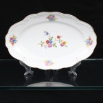 Porcelana Schmidt, Santa Catarina, Brasil (Fundada em 1945). Travessa rasa em porcelana nacional, c.1950/60, executado com elementos derivados do repertório Luís XV. Formato oval, borda ondulada contornada por friso em douradura, orla circundada por faixa de motvo fitomorfo relevado na própria pasta. Esmaltagem vítrea na cor branca, decoração floral policroma aplicada por decalcomania, constituída por ramagens e corolas em rosa, azul, vermelho, amarelo e folhagens verde. Marca da manufatura ao fundo. Medidas: Comp.: 28 x Larg.: 18 cm.