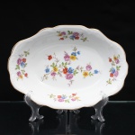 Porcelana Schmidt, Santa Catarina, Brasil (Fundada em 1945). Travessa funda em porcelana nacional, c.1950/60, executado com elementos derivados do repertório Luís XV. Formato oval, borda ondulada contornada por friso em douradura, orla circundada por faixa de motvo fitomorfo relevado na própria pasta. Esmaltagem vítrea na cor branca, decoração floral policroma aplicada por decalcomania, constituída por ramagens e corolas em rosa, azul, vermelho, amarelo e folhagens verde. Marca da manufatura ao fundo. Medidas: Alt.: 6 x Comp.: 28 x Larg.: 19,5 cm.