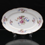 Porcelana Real, Mauá - SP(Fundada em 1943 por Fritz Erwin Schmidt). Belíssima travessa oval executada em porcelana nacional, circa 1960-1970. Esmaltagem vítrea na cor branca, ornamentação com motivos pictóricos policromados por processo de decalcomania fina inspirados no estilo "Deutsche Blumen", replicado nas reservas periféricas da orla, aba percorrida por gomos ondulares suaves, realçado com filetagem à ouro nas bordas e margens. Marca da manufatura no verso. Medidas: Comp.: x Larg.: cm.