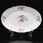 Porcelana Real, Mauá - SP(Fundada em 1943 por Fritz Erwin Schmidt). Belíssima travessa oval executada em porcelana nacional, circa 1960-1970. Esmaltagem vítrea na cor branca, ornamentação com motivos pictóricos policromados por processo de decalcomania fina inspirados no estilo "Deutsche Blumen", replicado nas reservas periféricas da orla, aba percorrida por gomos ondulares suaves, realçado com filetagem à ouro nas bordas e margens. Marca da manufatura no verso. Medidas: Comp.: x Larg.: cm.