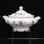 Porcelana Schmidt, Santa Catarina, Brasil (Fundada em 1945). Terrina coberta, para vegetais, em porcelana nacional, c.1950/60, executado com elementos derivados do repertório Luís XV. Bojo elipsoide assentado sobre pé moldurado de contorno recortado, alças laterais em arcos modelados com volutas e ressaltos, tampa abaulada encimada por pomo moldado com feitio de fruto sobre laçarote. Esmaltagem vítrea na cor branca, decoração floral policroma aplicada por decalcomania, constituída por ramagens e corolas em rosa, azul, vermelho, amarelo e folhagens verde., realçado por generosa filetagem em douradura nas bordas, pomo, base e alças laterais. Marca da manufatura ao fundo. Medidas: Alt.: 17 x Comp.: 27,5 x Larg.: 21 cm.