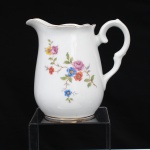 Porcelana Schmidt, Santa Catarina, Brasil (Fundada em 1945). Leiteira em porcelana nacional, c.1950/60, executado com elementos derivados do repertório Luís XV. Bojo oval alongado, borda recortada com vertedor pronunciado, asa lateral em voluta. Esmaltagem vítrea na cor branca, decoração floral policroma por decalcomania, constituída por ramagens e corolas em rosa, azul, vermelho, amarelo e folhagens verde, complementada por filetagem em dourada na base, borda, e alça. Marca da manufatura ao fundo. Medidas: Alt.: 14 x Comp.: 14,5 x Larg.: 9 cm.