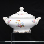 Porcelana Schmidt, Santa Catarina, Brasil (Fundada em 1945). Terrina coberta, para vegetais, em porcelana nacional, c.1950/60, executado com elementos derivados do repertório Luís XV. Bojo elipsoide assentado sobre pé moldurado de contorno recortado, alças laterais em arcos modelados com volutas e ressaltos, tampa abaulada encimada por pomo moldado com feitio de fruto sobre laçarote. Esmaltagem vítrea na cor branca, decoração floral policroma por decalcomania, distribuída pelo fundo e pelas paredes internas em buquês e ramagens com rosas, tulipa e flores miúdas, em rosa, azul, vermelho, amarelo e verde, realçado por generosa filetagem em douradura nas bordas, pomo, base e alças laterais. Marca da manufatura ao fundo. Medidas: Alt.: 18 x Comp.: 27 x Larg.: 22 cm.