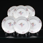 Porcelana Real, Mauá - SP (Fundada em 1943 por Fritz Erwin Schmidt). Conjunto de pratos para sobremesa em porcelana de pasta dura, fabricação nacional, circa 1960-1970. Esmaltagem vítrea na cor branca, centro dotado de ornamentação com motivos pictóricos policromados por processo de decalcomania fina inspirados no estilo Deutsche Blumen, completado com filetagem à ouro na borda e ao redor da caldeira, reservas na orla retratam um buquê clássico composto por rosa púrpura centralizada, violetas, miosótis e folhagens detalhadas em tons esmeralda, aba percorrida por gomos ondulares suaves. Marca da manufatura no verso. Composto de seis(06) unidades. Medida: Diam.: 18 cm.