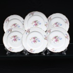 Porcelana Real, Mauá - SP (Fundada em 1943 por Fritz Erwin Schmidt). Conjunto de pratos rasos em porcelana de pasta dura, fabricação nacional, circa 1960-1970. Esmaltagem vítrea na cor branca, centro dotado de ornamentação com motivos pictóricos policromados por processo de decalcomania fina inspirados no estilo Deutsche Blumen, completado com filetagem à ouro na borda e ao redor da caldeira, reservas na orla retratam um buquê clássico composto por rosa púrpura centralizada, violetas, miosótis e folhagens detalhadas em tons esmeralda, aba percorrida por gomos ondulares suaves. Marca da manufatura no verso. Composto de seis(06) unidades. Medida: Diam.: 24 cm.