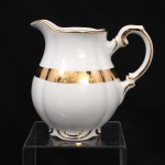 Porcelana Rio Branco / Cia. Brasileira de Vidros, Rio de Janeiro - RJ (fundada em 1947, ativa até 1967). Leiteira em porcelana nacional da década de 1950-60, executado ao estilo francês do período Luís XV. Bojo piriforme moldado com gomos em relevo, borda reocortada em arcos, tampa campaniforme encimada por pomo ovoide, bico vertedor ondulado e revirado para fora, asa alta em forma de voluta. em porcelana nacional. Esmaltagem vítrea na cor branca, com bordos de perfil ondulado, ornamentação constituída por faixa dourada com friso de ramagens florais estilizadas, complementada por filetes a ouro no bojo, borda e na alça. Marca da mnanufatura ao fundo. Medidas: Alt.: x Comp.: x Larg.: cm.