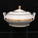 Porcelana Rio Branco / Cia. Brasileira de Vidros, Rio de Janeiro - RJ (fundada em 1947, ativa até 1967). Sopeira em porcelana nacional da década de 1950-60, executado ao estilo francês do período Luís XV. Bojo elipsoide moldado com gomos em relevo, assente sobre base circular, tampa abaulada encimada por pomo ovoide, alças laterais reviradas para cima. Esmaltagem vítrea na cor branca, com bordos de perfil ondulado, ornamentação constituída por faixa dourada com friso de ramagens florais estilizadas, complementada por filetes a ouro no bojo, borda e na alça. Marca da manufatura ao fundo. Medidas: Alt.: 15 x Comp.: 25 x Larg.: 22,5 cm.