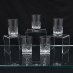 Conjunto de copos executados em meio cristal, diáfano e incolor, formato cilíndrico de base espessa, talhado com faixa geométrica composta por losangos facetados e cortes diagonais entrecruzados na porção inferior. Composto de cinco(05) unidades. Medidas. Altura  x Diâmetro:  cm.