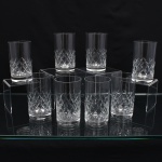 Conjunto de copos executados em meio cristal, diáfano e incolor, formato cilíndrico de base espessa, talhado com faixa geométrica composta por losangos facetados e cortes diagonais entrecruzados na porção inferior. Composto de oito(08) unidades. Medidas. Altura  x Diâmetro:  cm.