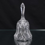 Sineta de mesa em cristal incolor prensado, corpo campaniforme de contorno lobulado, trabalhado em reservas ogivais alternadas com motivos geométricos, losangos reticulados e incisões dispostas radialmente. Haste alongada de seção facetada, integrada à campânula, culminando em remate lanceolado. Borda inferior recortada em sucessão de lóbulos. Decoração executada por lapidação, combinando planos polidos, sulcos e campos quadriculados. Medidas: Alt.: 17,5 x Diam.: 9cm.