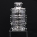 Pote com tampa em vidro prensado incolor, corpo cilíndrico constituído por três registros delimitados por frisos anulares, apresentando ornamentação de estrelas em relevo distribuídas pela superfície. Tampa de encaixe de perfil escalonado, rematada por pega tubular moldada e topo com textura granulada. Medidas: Alt.: 17 x Diam.: 10cm.