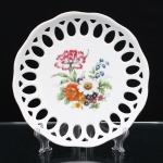 Porcelana Schmidt, Santa Catarina, Brasil (Fundada em 1945). Prato decorativo executado em porcelana nacional, década de 1960-70. Esmaltagem vítrea na cor branca, aba fenestrada com reservas lanceoladas e triangulares dispostas radialmente. Centro decorado com composição botânica policroma aplicada por decalcomania, representando cravo, margarida e demais flores, nas tonalidades vermelho-carmim, rosa, branco, laranja, amarelo, azul e violeta, entre folhagens verdes. Marca no verso: "Porcelana Schmidt - S. Catarina - Made in Brazil". Medida: Diam.: 19cm.
