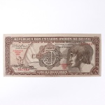 Numismática / Cédulas do Brasi. Cédula de 5 Cruzeiros, Estampa 3, Série 096, emitida pela República dos Estados Unidos do Brasil e impressa pela Casa da Moeda do Brasil no contexto da implantação do padrão monetário Cruzeiro, instituído em 1942 após a reforma que substituiu o sistema do réis. Anverso apresenta composição dominada por busto feminino alegórico representando a República, voltado à esquerda e inscrito em medalhão oval, tendo ao fundo vinheta marítima com embarcação à vela singrando mar agitado, elemento simbólico associado ao progresso econômico, à navegação e ao comércio. No campo central destaca-se elaborada cartela ornamental com o numeral 5 e a legenda CRUZEIROS, envolta por complexa trama de guilhochê de segurança característica da gravura calcográfica. Consta ainda a inscrição legal No Tesouro Nacional se pagará ao portador desta quantia de cinco cruzeiros, acompanhada das assinaturas fac-similares das autoridades monetárias e da indicação tipográfica Estampa 3 Série 096 impressa em vermelho. O reverso exibe vinheta naturalista representando vitória-régia (Victoria amazonica) em paisagem lacustre amazônica, com grandes folhas circulares flutuantes e flor central, enquadrada por moldura ornamental geométrica com os numerais 5 nos cantos e a legenda Estados Unidos do Brasil. Impressão calcográfica com fundos guilhochados de segurança e gravação em aço, padrão característico da produção da Casa da Moeda brasileira nas décadas de 19401950. Exemplar em estado aproximado MBC (Muito Bem Conservado), apresentando vincos de circulação, porém com papel íntegro e boa definição da gravura.