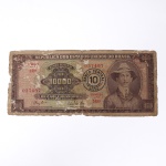 Numismática / Cédulas do Brasil. Cédula de 10.000 Cruzeiros com carimbo de sobrecarga de 10 Cruzeiros Novos, República dos Estados Unidos do Brasil, estampa 2ª, série 2494, impressa pela companhia britânica Thomas De La Rue & Company, Limited, circa 1967. O anverso apresenta fundo policromático em tons de sépia e marrom, ostentando medalhão circular à direita com o retrato de Alberto Santos-Dumont e medalhão losângico à esquerda com o valor de face original, modificado centralmente pelo carimbo circular de sobrecarga da transição monetária para o padrão Cruzeiro Novo (10 Cruzeiros Novos). O papel-moeda porta a numeração 037407. O reverso exibe composição em tom marrom-escuro com gravura calcográfica reproduzindo o voo do avião 14-Bis. O exemplar denota marcas severas de circulação, dobras, sujidade e desgaste perimetral com perda de papel nas bordas.
