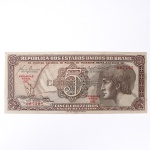 Numismática / Cédulas do Brasil. Cédula de 5 Cruzeiros, Estampa 3, Série 061, emitida pela República dos Estados Unidos do Brasil e impressa pela Casa da Moeda do Brasil no contexto da implantação do padrão monetário Cruzeiro, instituído em 1942 após a reforma que substituiu o sistema do réis. Anverso apresenta composição dominada por busto feminino alegórico representando a República, voltado à esquerda e inscrito em medalhão oval, tendo ao fundo vinheta marítima com embarcação à vela singrando mar agitado, elemento simbólico associado ao progresso econômico, à navegação e ao comércio. No campo central destaca-se elaborada cartela ornamental com o numeral 5 e a legenda CRUZEIROS, envolta por complexa trama de guilhochê de segurança característica da gravura calcográfica. Consta ainda a inscrição legal No Tesouro Nacional se pagará ao portador desta quantia de cinco cruzeiros, acompanhada das assinaturas fac-similares das autoridades monetárias e da indicação tipográfica Estampa 3 Série 061 impressa em vermelho. O reverso exibe vinheta naturalista representando vitória-régia (Victoria amazonica) em paisagem lacustre amazônica, com grandes folhas circulares flutuantes e flor central, enquadrada por moldura ornamental geométrica com os numerais 5 nos cantos e a legenda Estados Unidos do Brasil. Impressão calcográfica com fundos guilhochados de segurança e gravação em aço, padrão característico da produção da Casa da Moeda brasileira nas décadas de 1940-1950. Exemplar em estado aproximado MBC (Muito Bem Conservado), apresentando vincos de circulação, porém com papel íntegro e boa definição da gravura.
