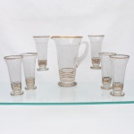 Conjunto para refresco em vidro, circa 19501960. Composto por uma(01) jarra e seis(06) copos, de linhas geométricas, com corpos obcônicos sobre bases circulares. Jarra provida de bico vertedor e asa aplicada em vidro maciço. Ornamentação executada por filetes verticais e faixas horizontais, complementada por frisos dourados nas bordas e bases. Copos de perfil alongado, com abertura alargada e pé circular. Medidas jarra: Alt.: 25 x Comp.: 21 x Larg.: 16cmMedidas copo: Alt.: 17 x Diam.: 8,5cm.