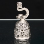 Sineta de mesa confeccionada em metal prateado, corpo campaniforme vazado, ornamentado em relevo com motivos fitomórficos e representações dos quatro Evangelistas, identificados pelas inscrições latinas S. Johannes, S. Mathevs, S. Lvcas e S. Marcvs. Empunhadura zoomórfica em forma de peixe estilizado, de corpo escamado e cauda em leque. Badalo interno de formato esférico. Medidas: Alt.: 9,5 x Diam.: 5CM.
