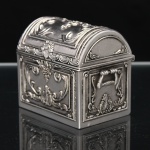 Caixa porta-objetos em forma de baú executado em metal prateado, corpo retangular com tampa abaulada articulada e alças laterais móveis. Superfícies ornamentadas em relevo com volutas, palmetas, concheados, frisos perlados e motivos fitomórficos, em repertório de inspiração neorrococó. Interior liso. Medidas: Alt.: 8 x Comp.: 9 x Larg.: 6cm.