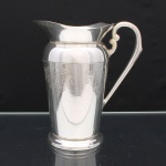 EBERLE. Elegante jarra confeccionada em metal espessurado à prata, bojo balaústre ricamente lavrada com elementos vegetalistas, assente sobre base circular repuxada em degraus, asa lateral em volutas, boda ondulada e revirada para fora, interior dotado de barra retentora de gelo. Marca da manufatura ao fundo. Med.: Alt. 22 x Comp. 20 x Larg. 13 cm.