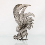 Escultura animalista representando galo, executada em metal prateado. Figura executada por fundição, com plumagem cinzelada e gravada, asas parcialmente abertas e cauda composta por longas retrizes arqueadas. Cabeça inclinada, bico entreaberto, crista recortada, pernas com esporões e pés providos de garras. Medidas: Alt.: 26,5 x Comp.: 16 x Larg.: 17cm.