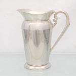EBERLE. Elegante jarra confeccionada em metal espessurado à prata, bojo balaústre ricamente lavrada com elementos vegetalistas, assente sobre base circular repuxada em degraus, asa lateral em volutas, boda ondulada e revirada para fora, interior dotado de barra retentora de gelo. Marca da manufatura ao fundo. Med.: Alt. 22 x Comp. 20 x Larg. 13 cm.