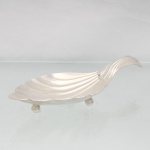 Elegante petisqueira confeccionada em metal espessurado à prata, executada na forma de concha, interior côncavo repuxado com sulcos ondulados, alça lateral curvada para dentro, assente sobre três pés esféricos. Med.: Alt.: 4 x Comp.: 20 x Larg.  9cm.