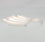 Elegante petisqueira confeccionada em metal espessurado à prata, executada na forma de concha, interior côncavo repuxado com sulcos ondulados, alça lateral curvada para dentro, assente sobre três pés esféricos. Med.: Alt.: 4,5 x Comp.: 20 x Larg.: 9cm.