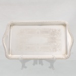Elegante travessa retangular confeccionada em metal espessurado a prata, centro ligeiramente rebaixado, ricamente lavrada com composição de motivo vegetalista, borda contornada por barrado relevado, extremos com abas onduladas integradas. Medidas: Comp.: 36 x Larg.: 20cm.